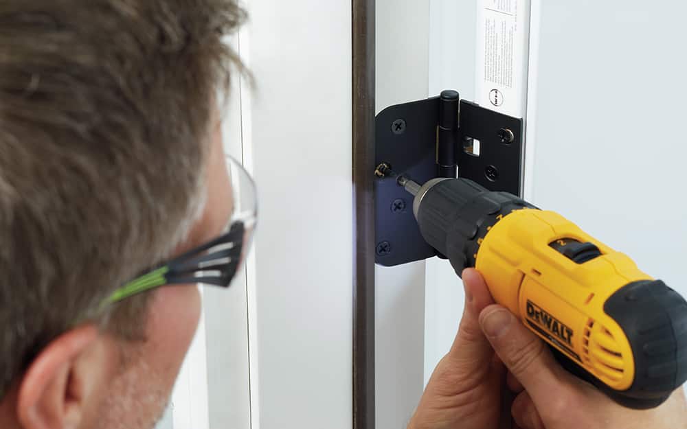 A man drilling hinges into a new entry door inside the home. A man drilling hinges into a new entry door inside the home.