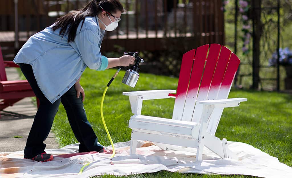 A woman using a paint sprayer to paint an an Adirondack chair. A woman using a paint sprayer to paint an an Adirondack chair.