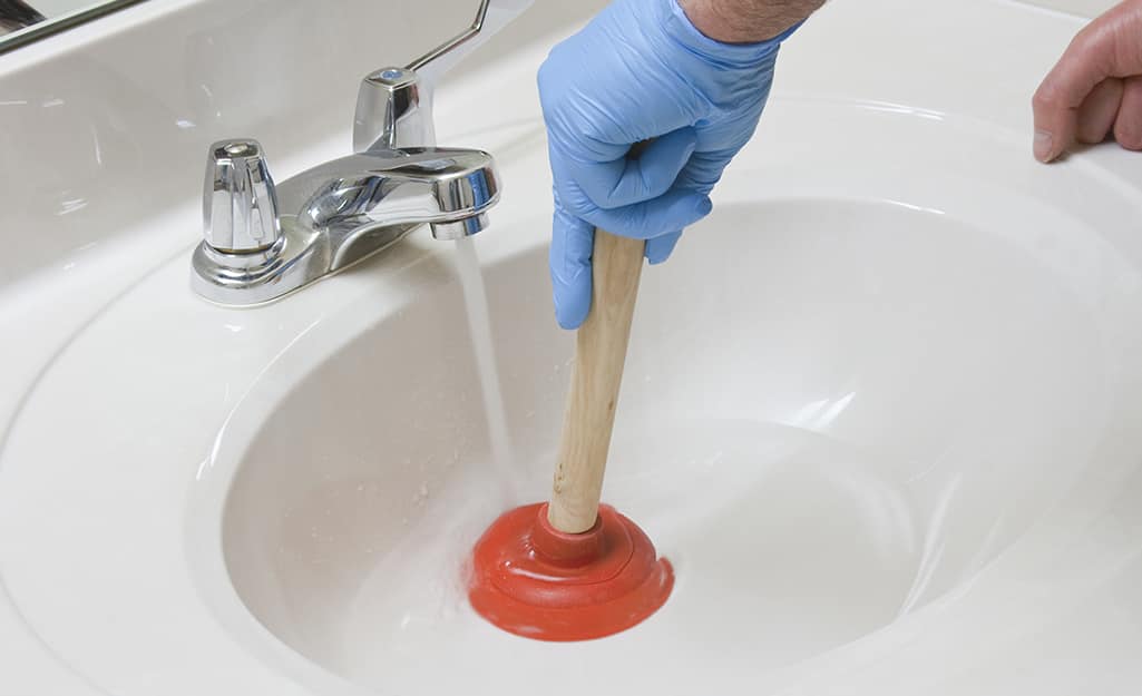 Someone wearing a blue glove and using a sink plunger under running water to unclog a sink. Someone wearing a blue glove and using a sink plunger under running water to unclog a sink.