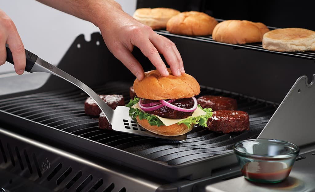 A person uses a metal spatula to lift a burger from a grate. A person uses a metal spatula to lift a burger from a grate.