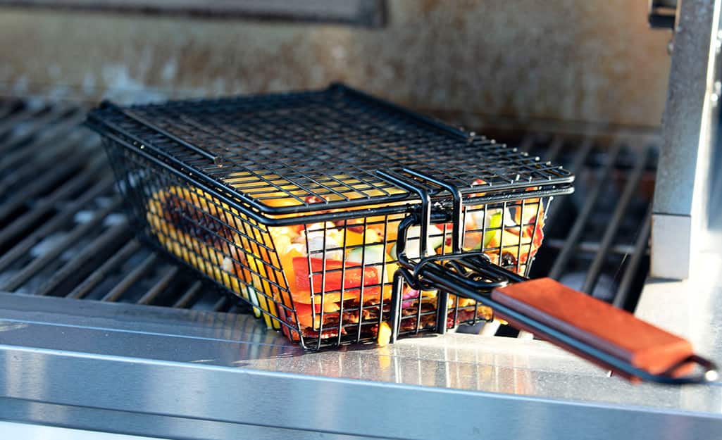 A long-handled grilling basket holds vegetables on a grill. A long-handled grilling basket holds vegetables on a grill.