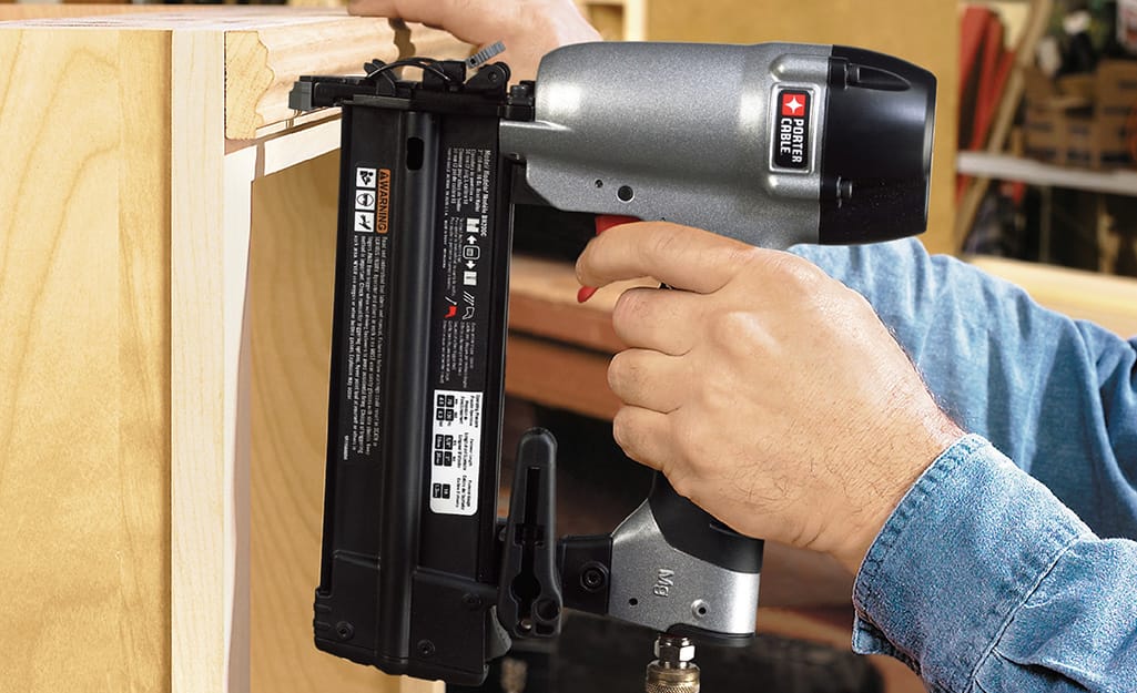 Someone using a finish nailer to fasten moulding along the top of a bookshelf. Someone using a finish nailer to fasten moulding along the top of a bookshelf.