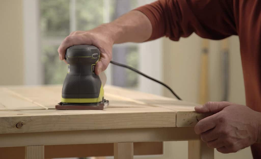 A man using a power sander to sand the top of a DIY coffee table. A man using a power sander to sand the top of a DIY coffee table.