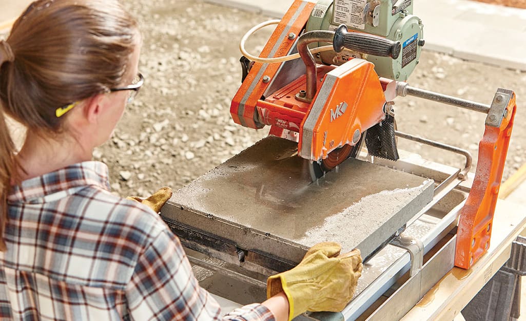 A woman in safety eyewear and work gloves using a concrete saw to cut pavers for a walkway. A woman in safety eyewear and work gloves using a concrete saw to cut pavers for a walkway.