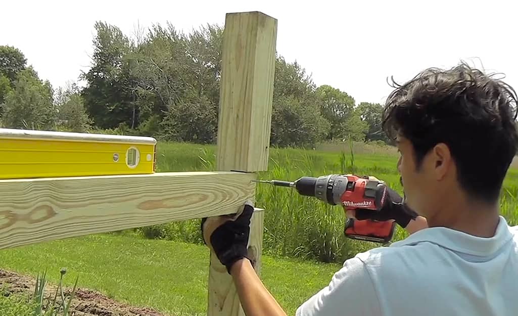 A person securing the top fence rail to the fence post with a drill and screws. A person securing the top fence rail to the fence post with a drill and screws.