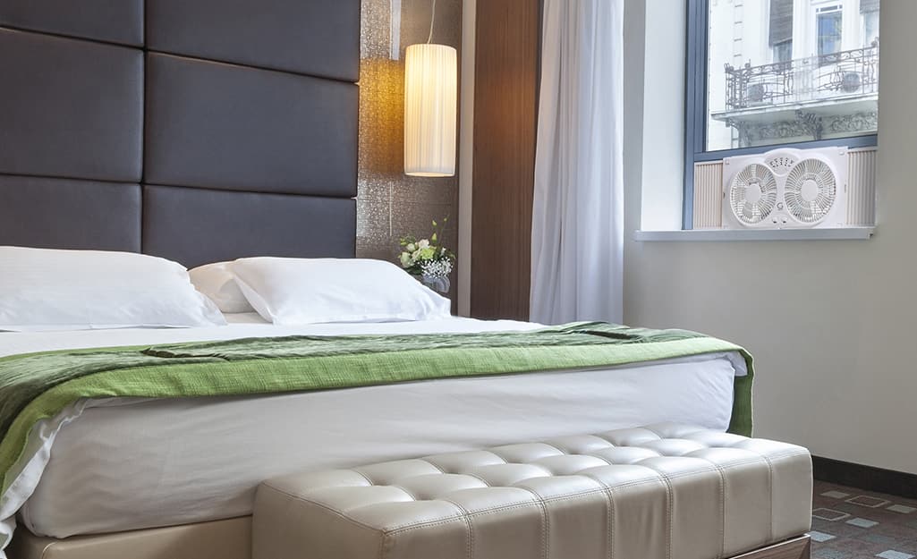 A bedroom with a window fan in the window. A bedroom with a window fan in the window.