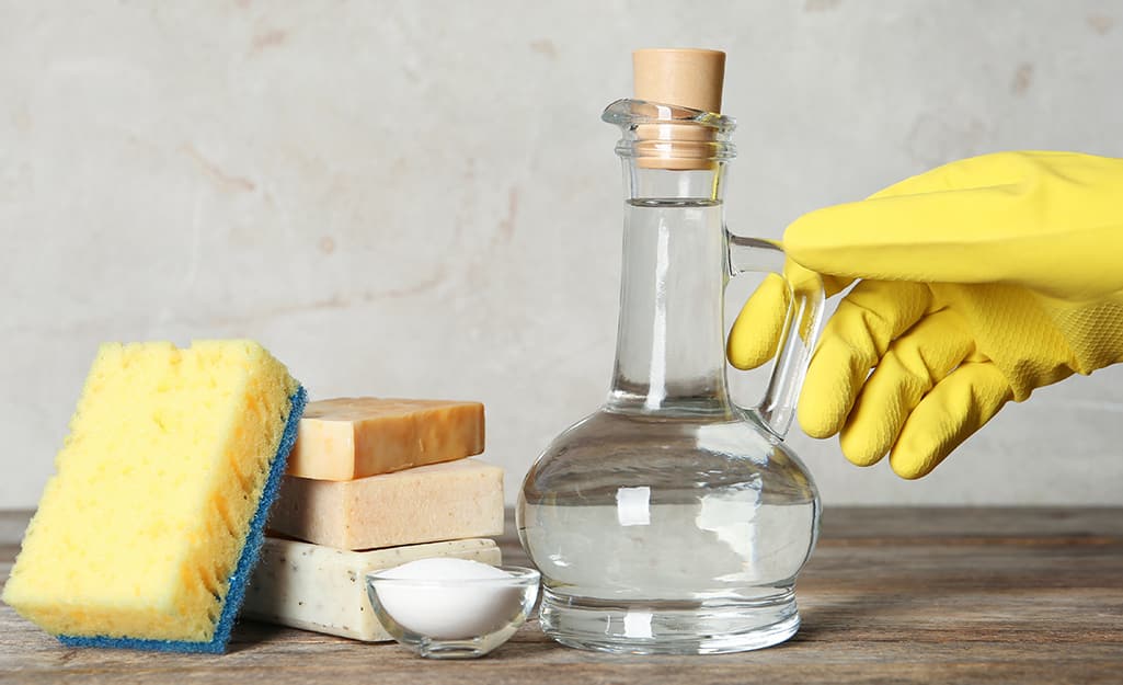 A hand in a dishwashing glove reaches for a clear bottle of vinegar that sits next to baking soda, soap and a sponge. A hand in a dishwashing glove reaches for a clear bottle of vinegar that sits next to baking soda, soap and a sponge.