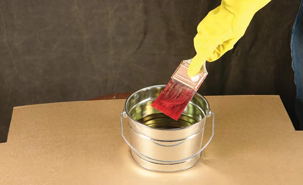 A person wearing kitchen gloves cleans a paint brush in a small container of solvent. A person wearing kitchen gloves cleans a paint brush in a small container of solvent.