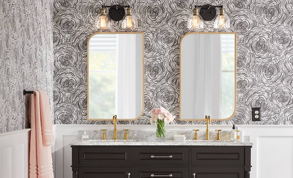 A bathroom with abstract wallpaper and matching mirrors with lamps hung over a dark vanity with gold finished faucets.
