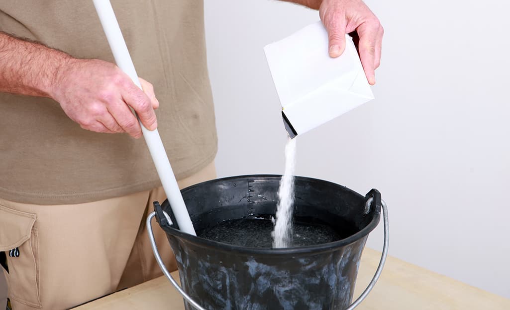A man pours an unlabeled box of grout powder into a bucket. A man pours an unlabeled box of grout powder into a bucket.