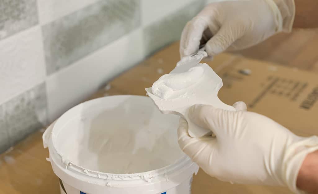 Gloved hands scoop epoxy grout from a bucket with two scrapers. Gloved hands scoop epoxy grout from a bucket with two scrapers.