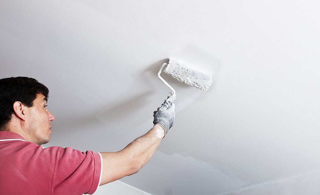 A man begins to paint the ceiling white using a roller. A man begins to paint the ceiling white using a roller.