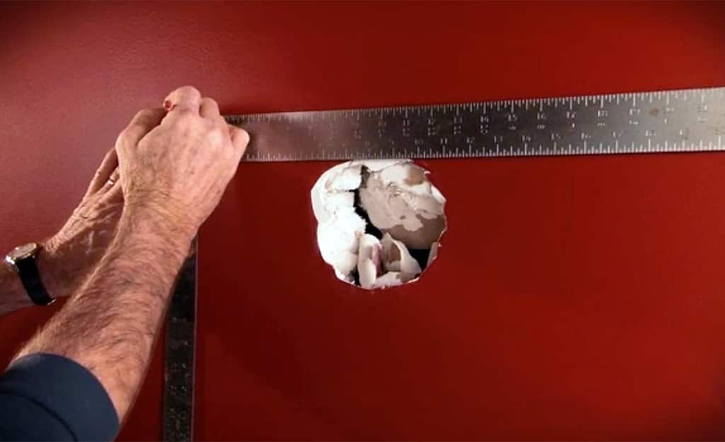 A person using a carpenter's square to measure a large hole in the wall. A person using a carpenter's square to measure a large hole in the wall.