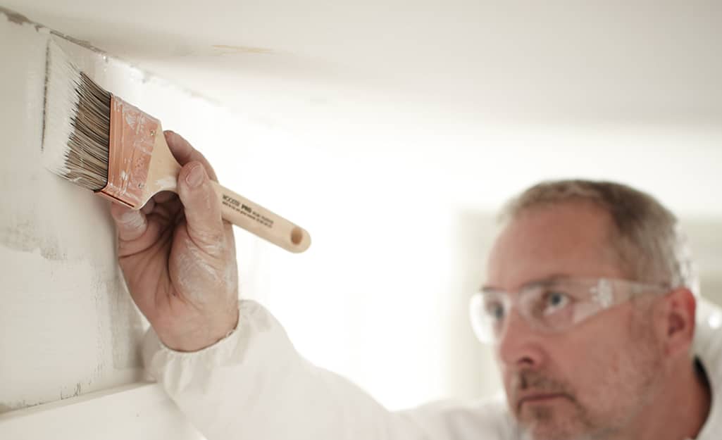A person using a paint brush to apply primer on a wall at the edge of the ceiling. A person using a paint brush to apply primer on a wall at the edge of the ceiling.