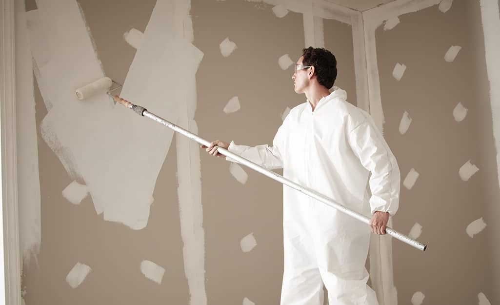 A person using a paint roller on an extension pole to apply primer to an unfinished wall. A person using a paint roller on an extension pole to apply primer to an unfinished wall.