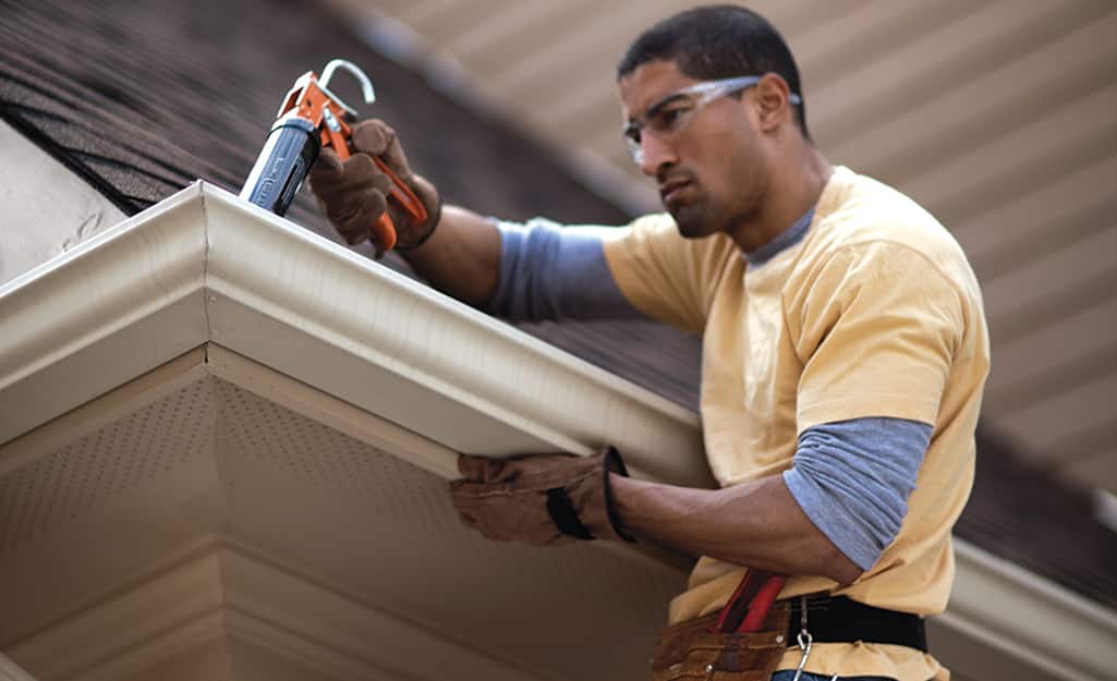 A person adding plastic roofing cement around white gutters on a roof's edge. A person adding plastic roofing cement around white gutters on a roof's edge.