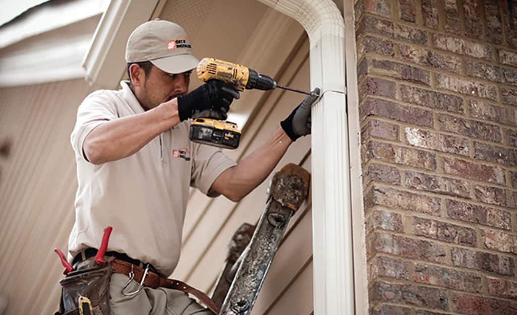 A person using a drill to reattach a downspout to the side of a house. A person using a drill to reattach a downspout to the side of a house.