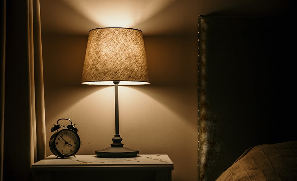 A rewired lamp on a bedside table illuminates a vintage alarm clock and bed. A rewired lamp on a bedside table illuminates a vintage alarm clock and bed.