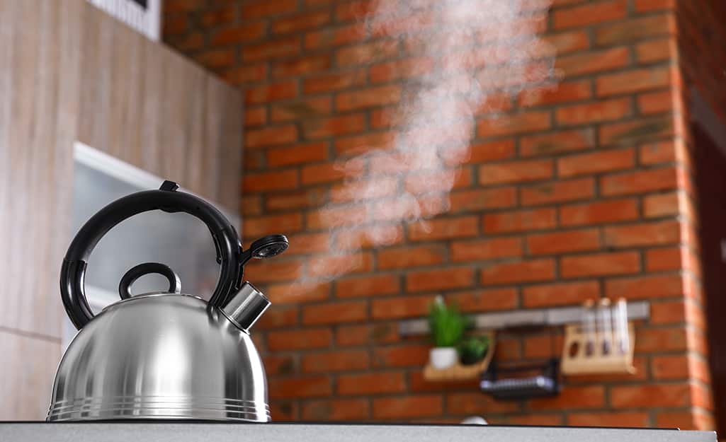 A silver kettle boils on a stove and its steam is visible against a brick wall A silver kettle boils on a stove and its steam is visible against a brick wall