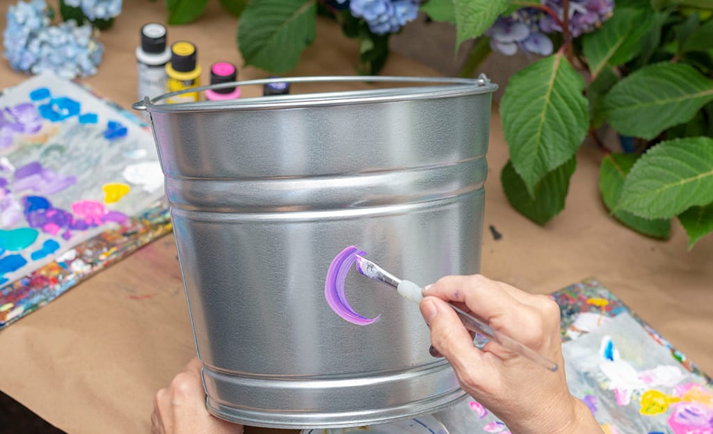 A person applying purple acrylic paint onto a galvanized bucket. A person applying purple acrylic paint onto a galvanized bucket.