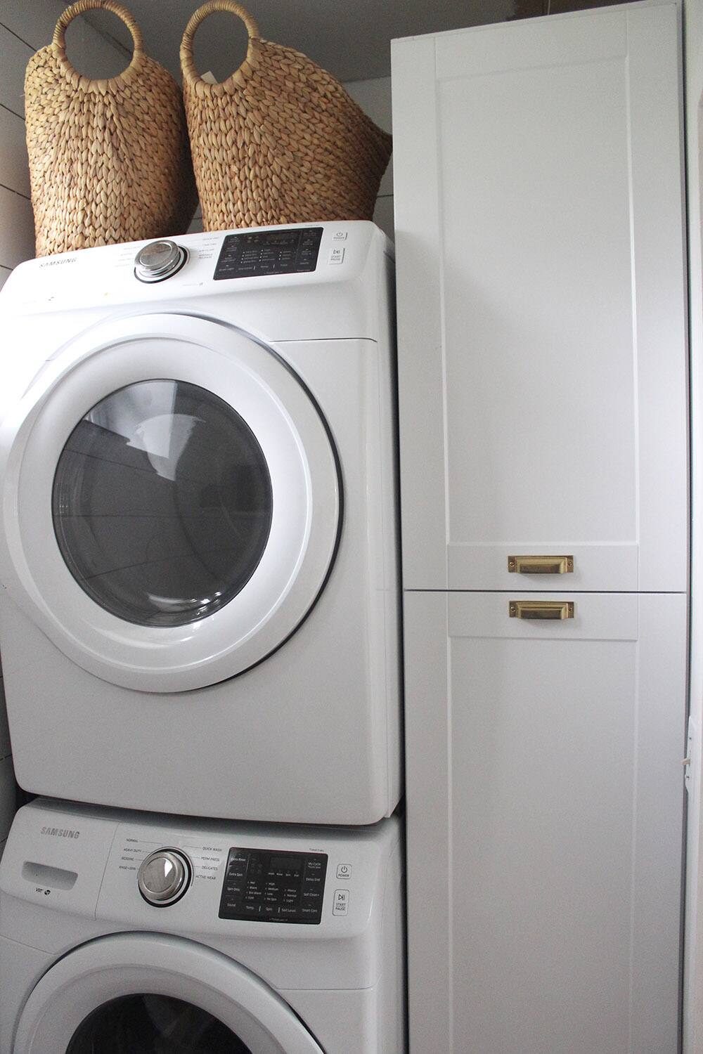 A laundry room with a washer, dryer, white cabinets with gold hardware, and woven baskets.