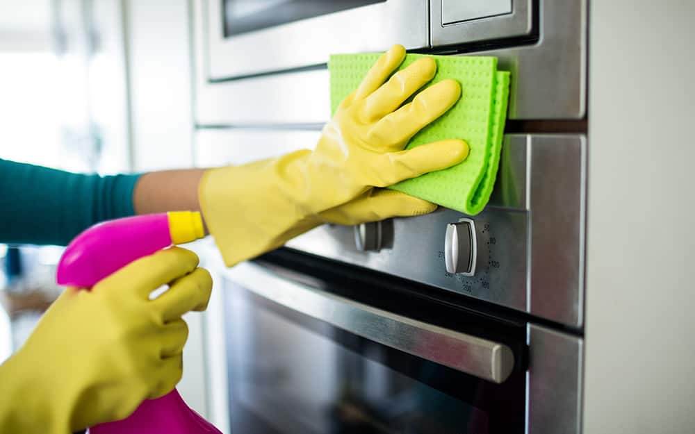 A person wearing gloves and cleaning the exterior of a wall oven with a sponge and spray bottle A person wearing gloves and cleaning the exterior of a wall oven with a sponge and spray bottle