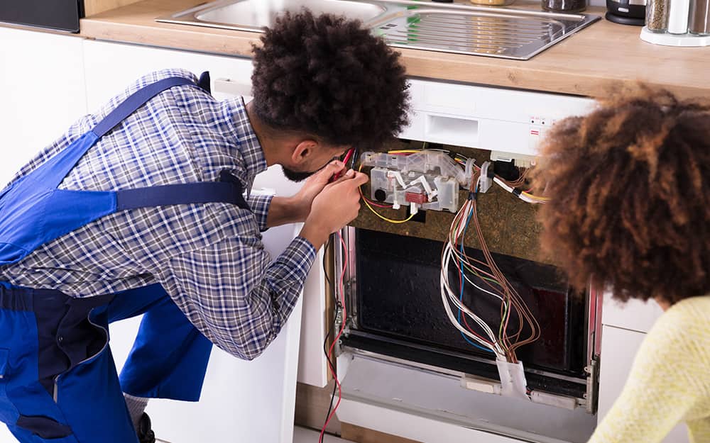 Two people look at the electrical components of a dishwasher. Two people look at the electrical components of a dishwasher.