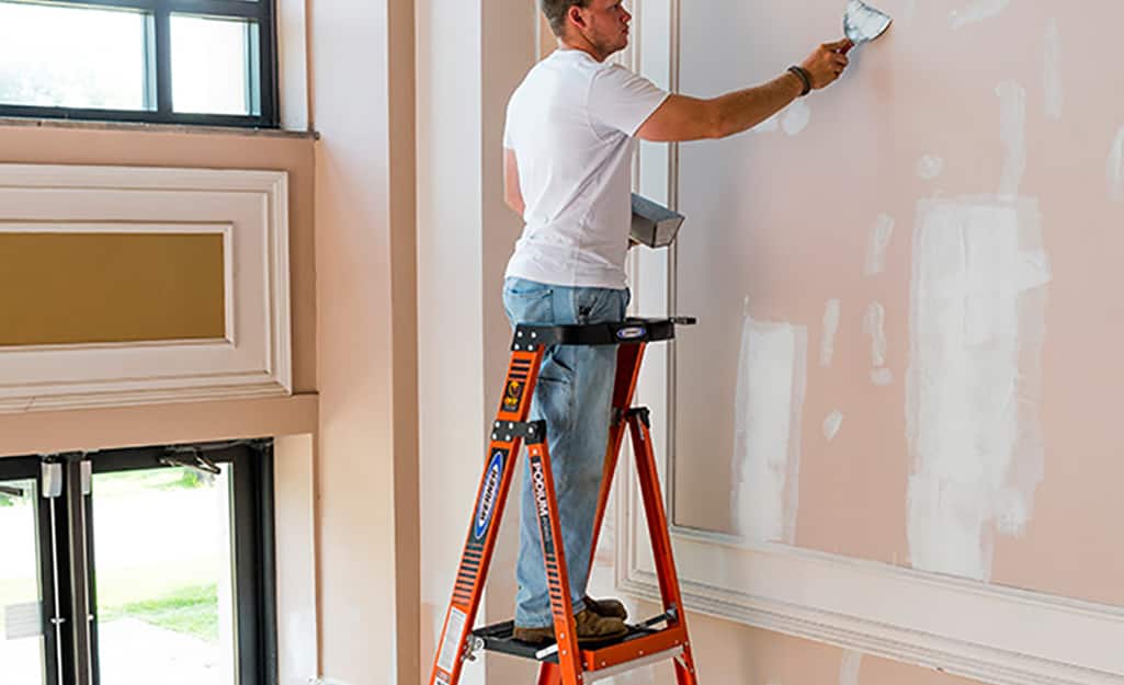 A man patching a wall while standing safely on a platform ladder. A man patching a wall while standing safely on a platform ladder.