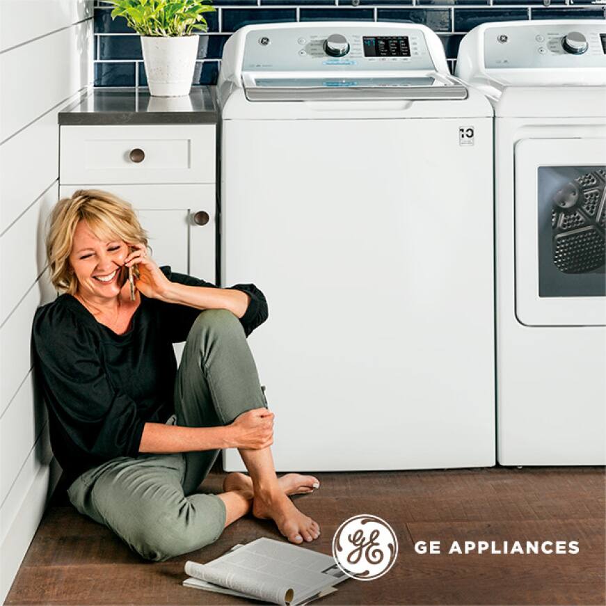 Woman sitting on the ground next to her GE combo washer and dryer set while talking on the phone and reading a magazine