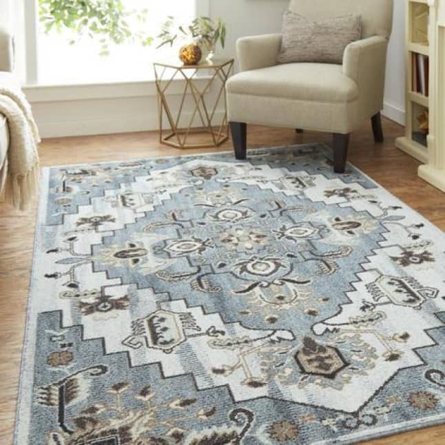 A blue hued, tribal designed rug sits in the center of a room with an armchair sitting in the top-right part of the image next to a fireplace.